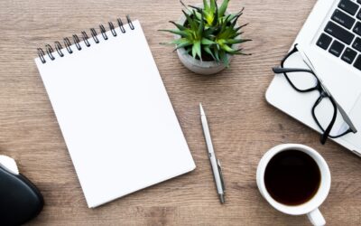 An open notebook, surrounded by a grey pen, a succulent plant, a mug full of coffee, and a pair of glasses resting on the bottom left corner of an open Apple laptop.